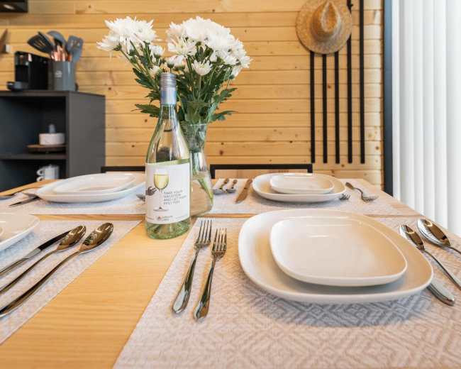 A dining table is set with white plates, cutlery, and a bottle of wine, accompanied by a vase of flowers and a straw hat in the background.