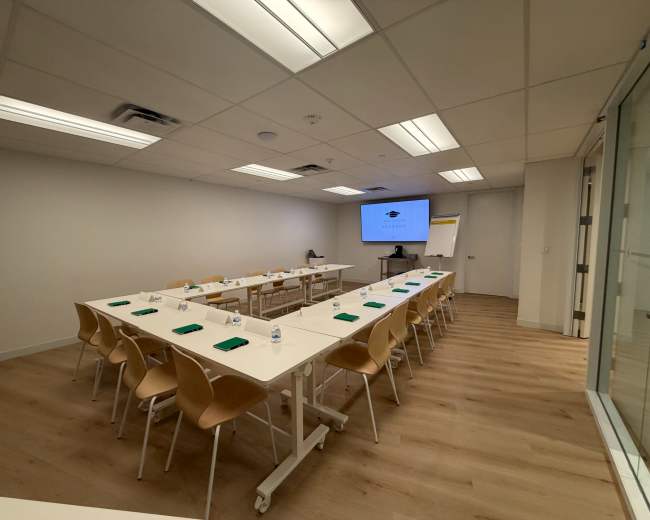 The image shows a modern conference room with a long table arranged with chairs, water bottles, and notepads, along with a presentation screen at one end.