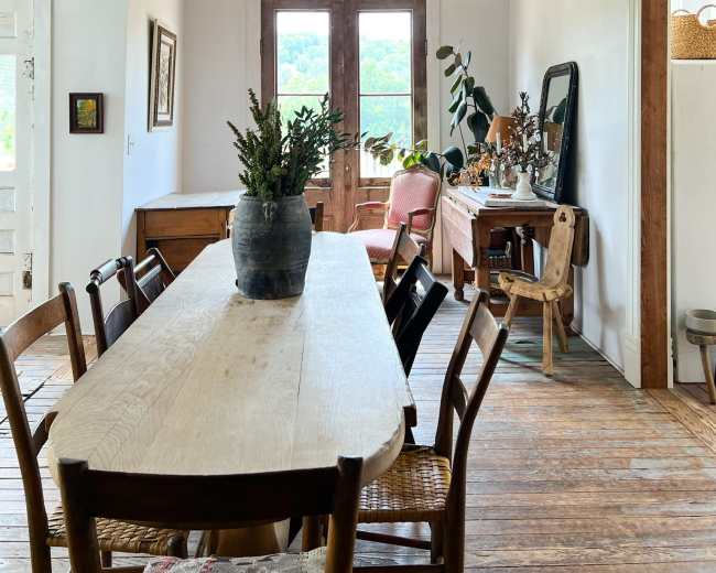 A long wooden dining table is centered in a room with a whitewashed wall, featuring a door that leads to a view of greenery outside.