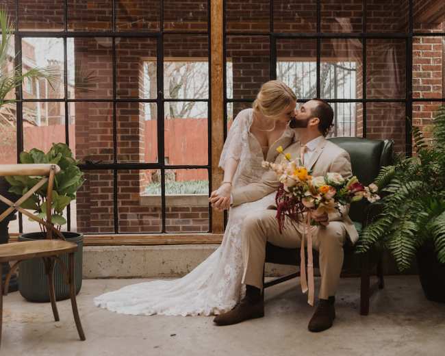 A bride and groom sit together in a plant-filled room, with the bride holding the groom's face as he holds a bouquet of flowers.