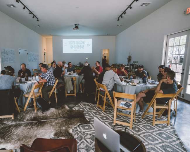 A group of approximately 30 people is seated around several tables in a well-lit meeting room, engaged in discussion with materials and laptops in front of them.