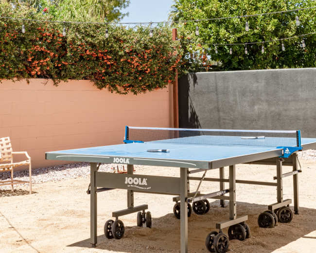 A ping pong table is set up on a sandy area surrounded by a pink wall and greenery under a clear blue sky.