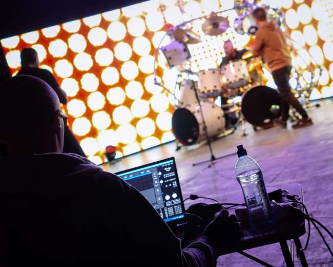 A person sits at a laptop in the foreground, while a drummer plays on stage in front of a backdrop of bright, circular lights.