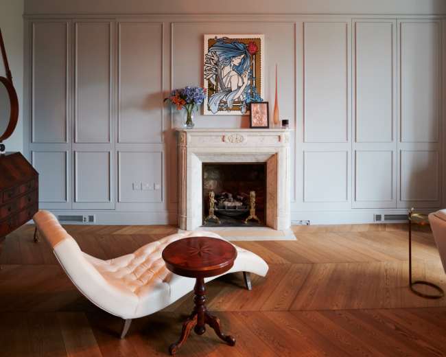 The room features a modern design with a white lounge chair, a small round table, a fireplace with a framed art piece above it, and pendant lights hanging from the ceiling.