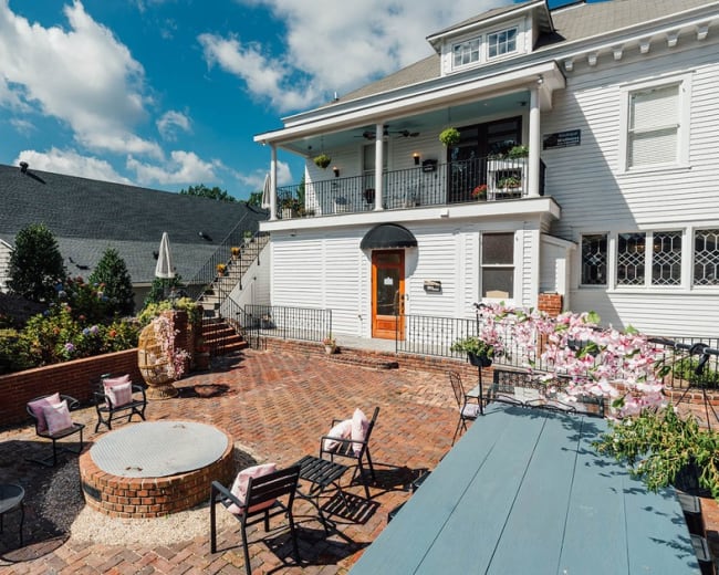A brick patio with seating and a fire pit is situated outside a large white house, featuring a balcony and landscaped garden.
