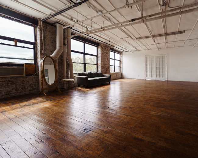 A spacious, minimally furnished room with wooden floors, large windows, and a couch, featuring exposed brick walls and a mirror.