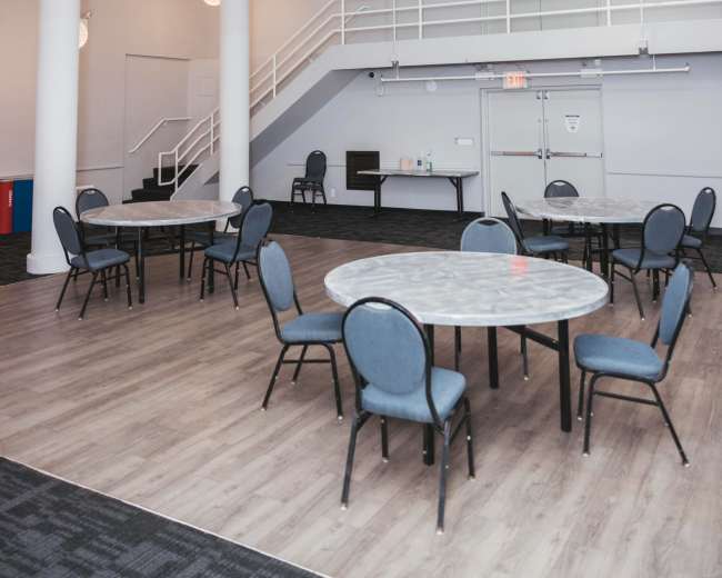 The image shows an empty event space with several round tables and chairs, modern light fixtures hanging from the ceiling, and a staircase in the background.