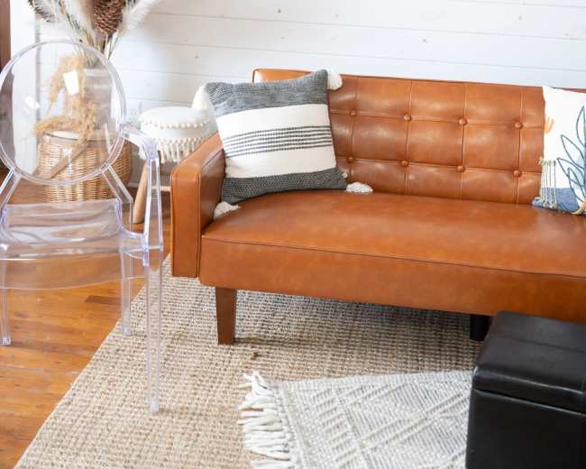 A brown leather sofa with tufted detailing sits next to a clear acrylic chair and decorative plants in a room with white walls.