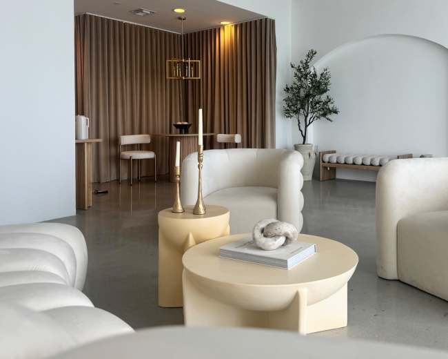 The image shows a modern, minimalist lounge area with white upholstered chairs and a light-colored coffee table, surrounded by beige curtains and a simple decor.