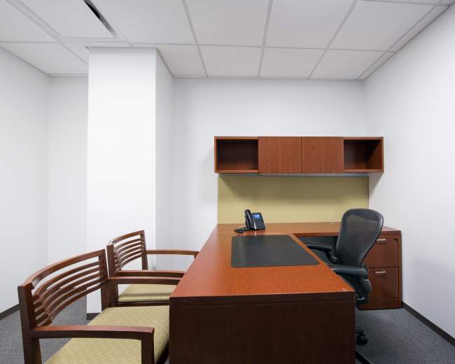 A minimalistic office space featuring a wooden desk, an office chair, and two wooden chairs against a plain wall.