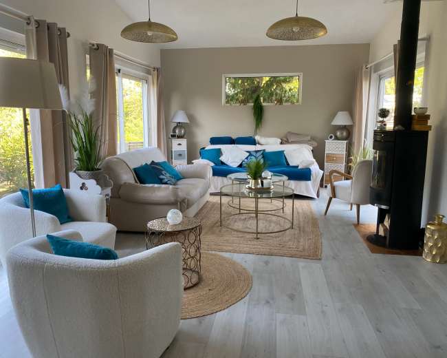 The living room features a light-colored sofa, a round coffee table, and various plants, with large windows letting in natural light.