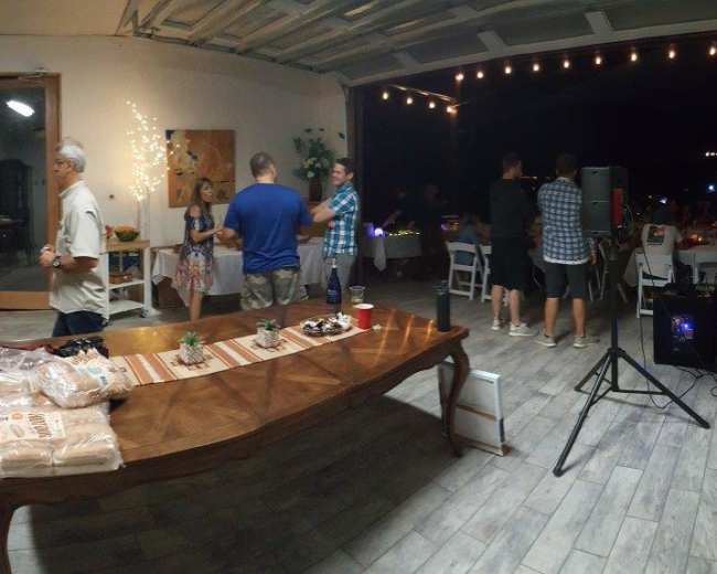 A group of people gather in a spacious kitchen and dining area, with a large wooden table set with food and drinks, while others enjoy conversation and music.