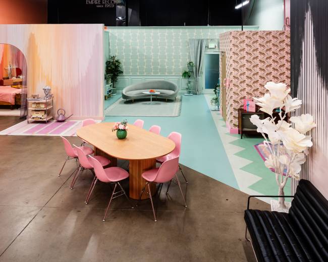 The image features a vibrant indoor scene with distinct pink and teal decor, including a dining table surrounded by pink chairs and various stylized room sections in the background.