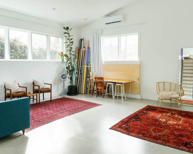 A bright, spacious room features a teal sofa, wooden chairs, a table, and decorative rugs against a backdrop of large windows and light-colored walls.