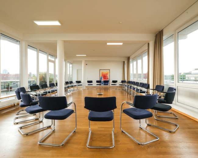 The image shows a spacious room arranged with multiple chairs in a circular formation around a central area, surrounded by large windows that allow natural light.