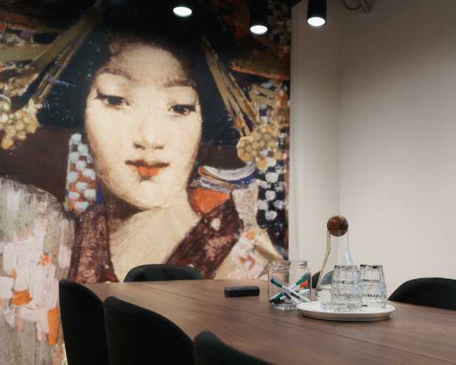 A conference room features a wooden table with glasses and a glass decanter, set against a mural of a woman in traditional attire.