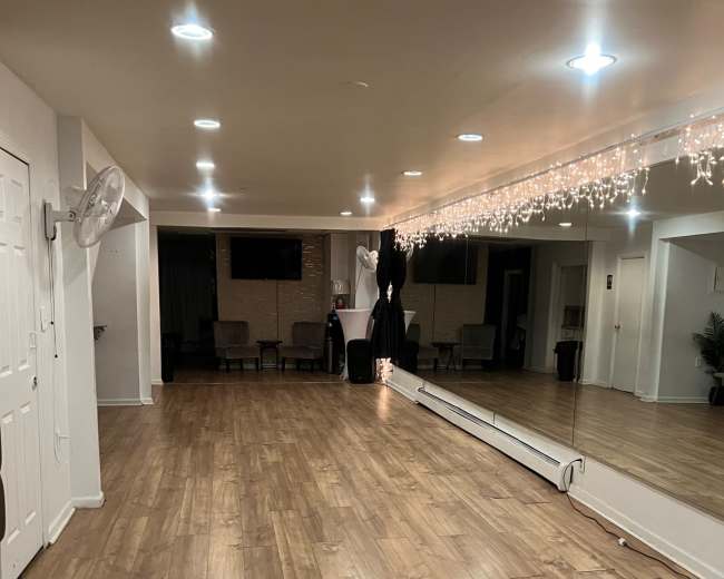 The image shows a spacious indoor area with wooden flooring, bright ceiling lights, a large mirror on one wall, and a few pieces of furniture.