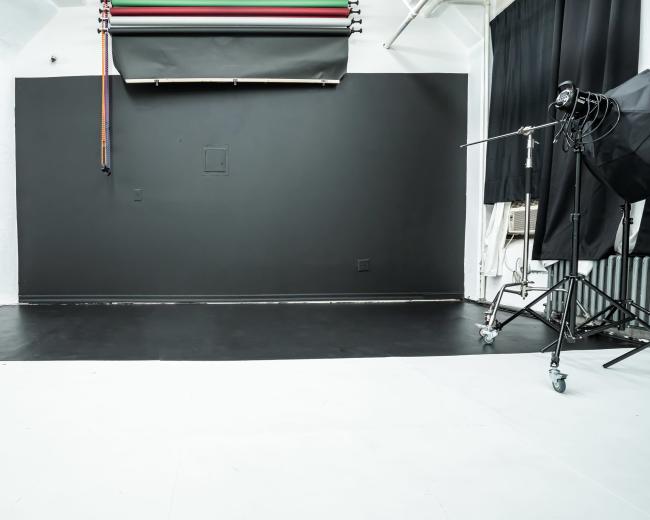 A photography studio with a black backdrop and a light stand on one side, along with a section of white floor.