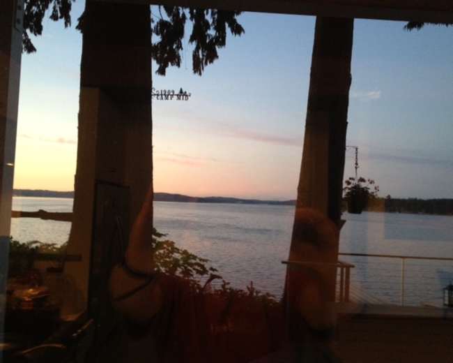 The image shows a tranquil waterfront view at sunset, with trees framing the scene and reflections visible on the glass.