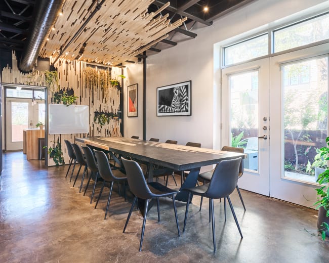 A large conference room features a wooden table surrounded by black chairs, with natural light streaming in through two doorways and decorative plants along the walls.