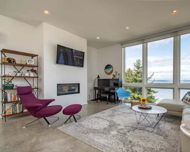 The image shows a modern living room featuring a purple chair, a white coffee table with a sunflower, a sofa with decorative pillows, a piano, and large windows offering a view of the outdoors.