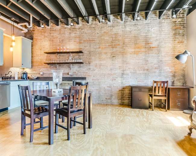 The image shows a modern kitchen and dining area with exposed brick walls, a wooden dining table surrounded by chairs, and a small desk in the corner.