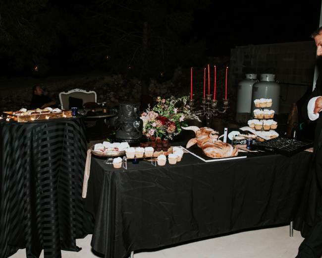 A table is set outdoors featuring an assortment of desserts, including cupcakes and pastries, alongside a loaf of bread and decorative floral arrangements, with dim lighting and a few seated individuals in the background.