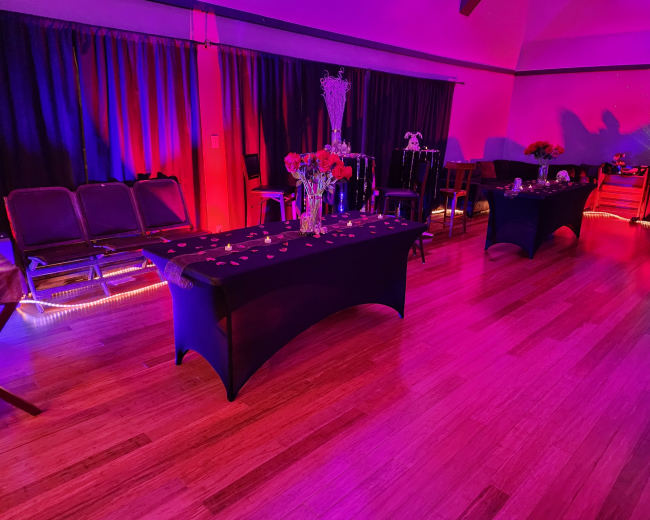 The scene shows a dimly lit event space with decorated tables, chairs lined against the wall, and purple and pink lighting creating a festive atmosphere.