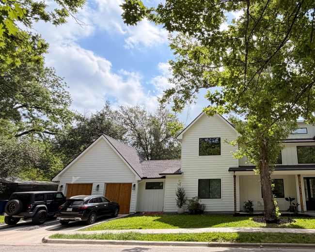 A two-story, modern farmhouse-style house with white exterior and a large front porch sits among trees on a residential street.