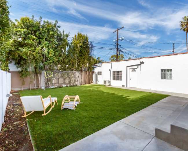 A spacious backyard features a green lawn, a seating area with two chairs, and a concrete patio adjacent to a white house.
