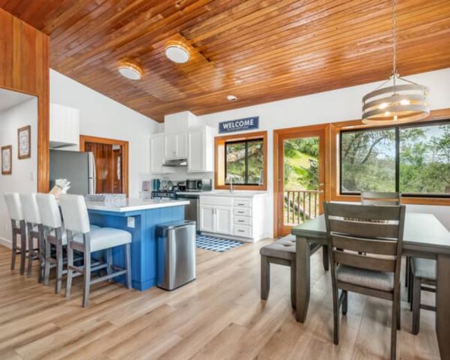 A modern kitchen and dining area features a wooden ceiling, white cabinetry, and a blue island surrounded by bar stools.