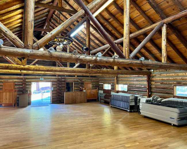 The interior of a spacious log cabin features exposed wooden beams and stacked logs, with folding tables stacked against the wall and large windows allowing natural light to enter.