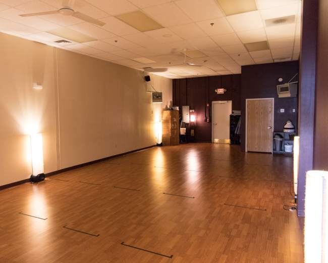 A spacious room with wooden flooring, lit by soft lights at either end, and a closed door on one side.