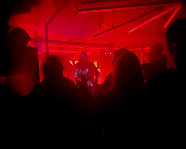 A DJ performs on stage in a dimly lit room with red lighting, while silhouettes of the audience are visible in the foreground.