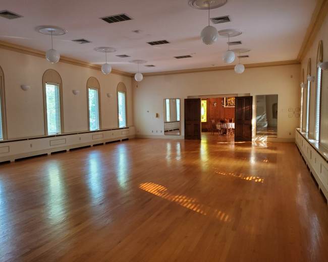 A spacious, well-lit room with wooden flooring, large windows, and a mirrored wall.