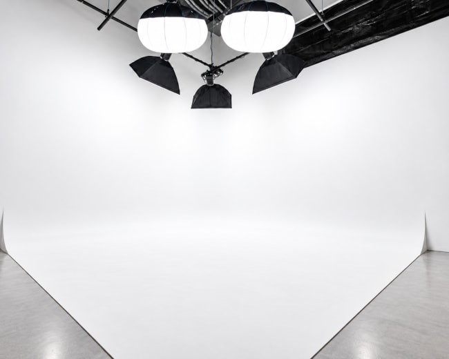 The image shows a spacious photography studio with a seamless white backdrop and large overhead lighting equipment.