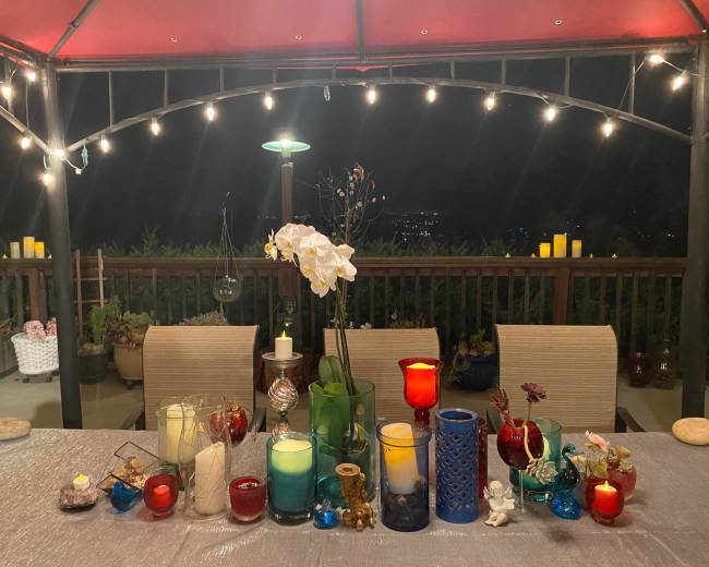 A display of colorful candles and decorative items is arranged on a table beneath a canopy, with a view of twinkling lights in the background.