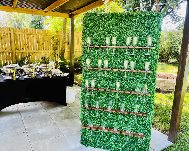 A green wall covered in foliage decorations stands next to a table with food serving trays in an outdoor setting.