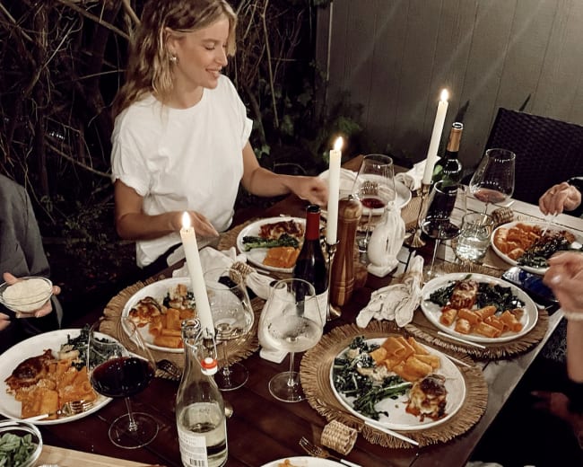 A group of people enjoys a dinner spread featuring various dishes, wine, and candles on a wooden table outdoors.