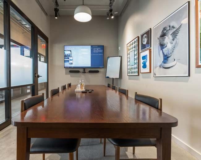 A large wooden conference table is set in a well-lit meeting room, featuring a television screen on the wall and several framed artworks.
