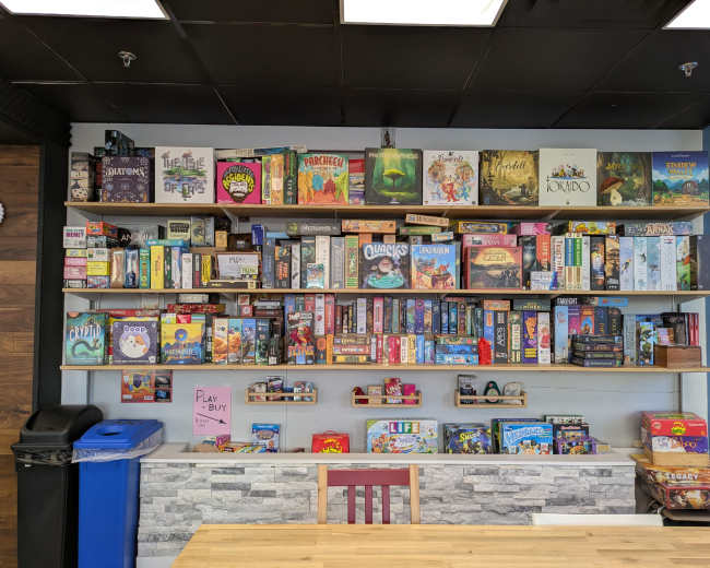A well-organized shelf displays a wide variety of board games in a cafe setting, with a table and trash bin visible below.