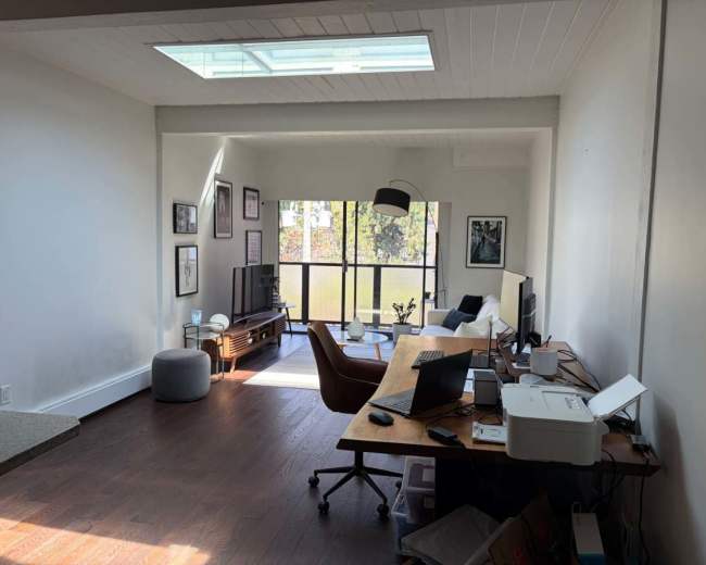 The image shows a spacious interior with a wooden floor, featuring a desk with a computer and printer, a comfortable chair, and a large window that provides natural light.