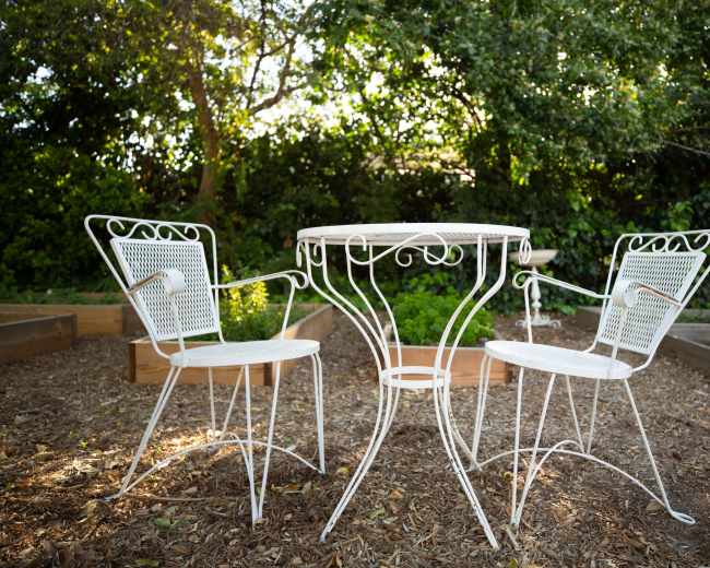 A white metal table with two matching chairs is positioned on mulch in a garden surrounded by greenery.