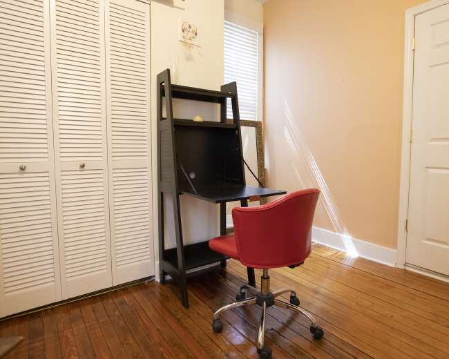 A black desk with a red chair is positioned against a wall in a room with wooden floors and a door.
