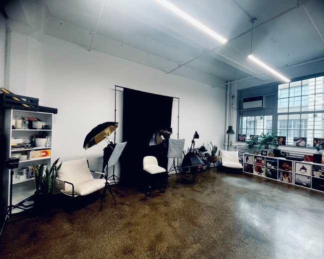The image shows a well-lit studio space featuring three photography lights on stands, a black backdrop, two white chairs, and shelves filled with various items.