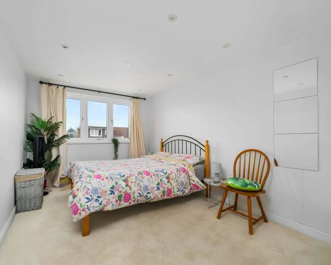 A simple bedroom features a bed with a floral blanket, a wooden chair, and a small storage basket, with a window allowing natural light to enter the space.