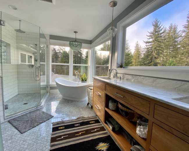 The bathroom features a glass shower, a freestanding bathtub, a wooden vanity with open shelving, and large windows overlooking a forest.