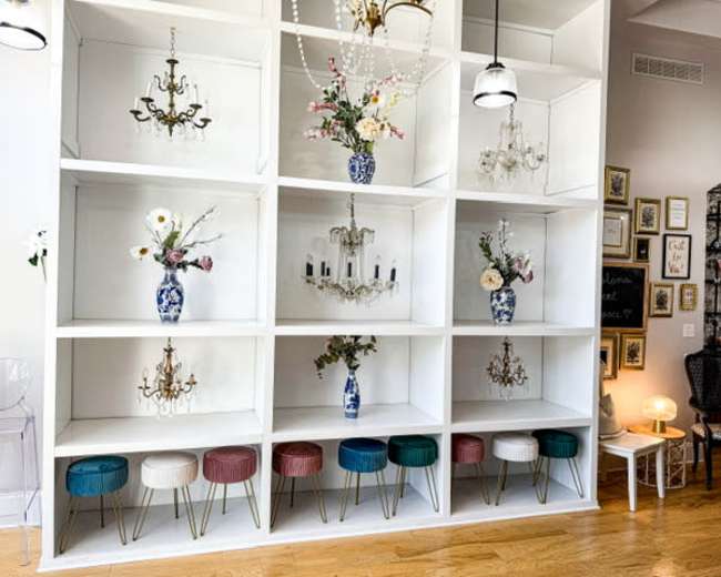 The image shows a white shelving unit with decorated niches featuring vases, floral arrangements, and small chandeliers, accompanied by colorful stools below.