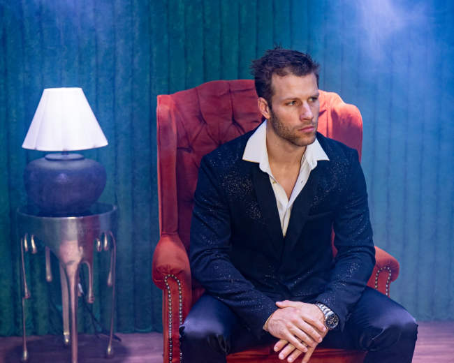 A man in a stylish black suit sits confidently in a red velvet armchair, surrounded by a smoky atmosphere and a lamp beside him.