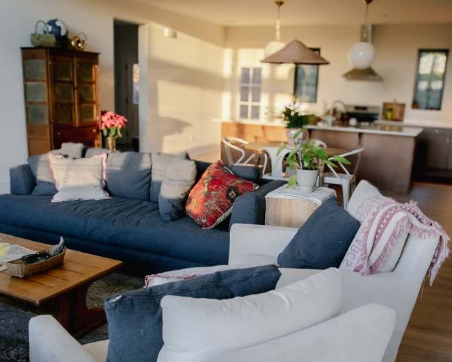 The image shows a modern living room with a large blue sofa, white armchairs, a wooden coffee table, and a kitchen area visible in the background.